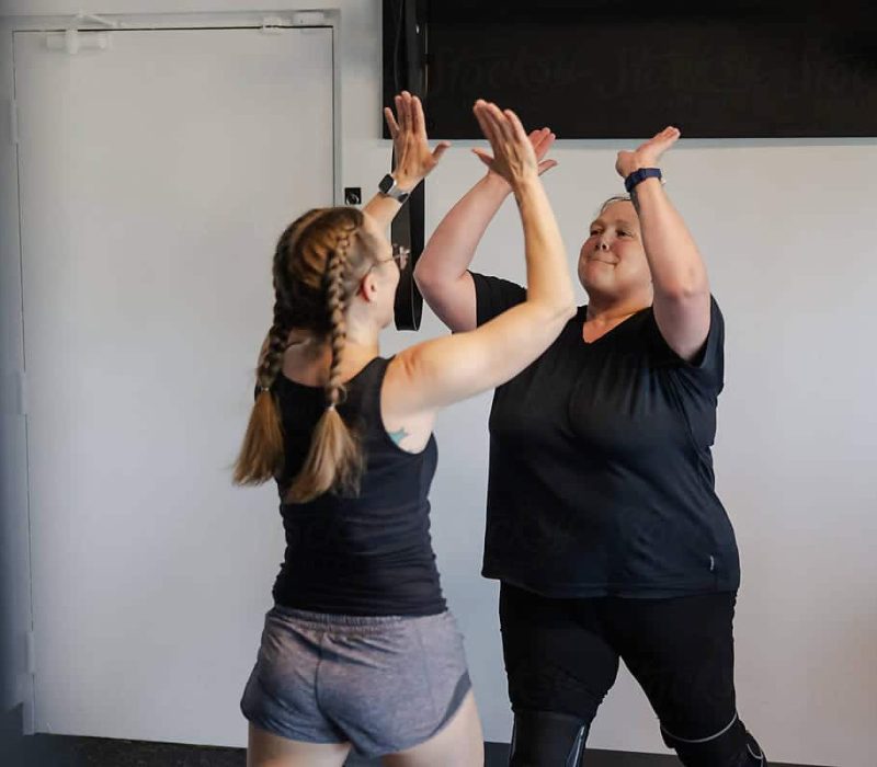 Two women in a gym celebrate a successful workout with a high five, showing determination, joy, and teamwork.
